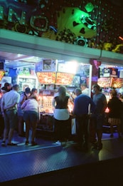 A group of people standing in front of an illuminated amusement arcade, engaging with various gaming machines. The environment is bustling with activity, and colorful lights create a vibrant atmosphere. The ceiling is decorated with large, playful images of dice and other game-related graphics, enhancing the lively mood.