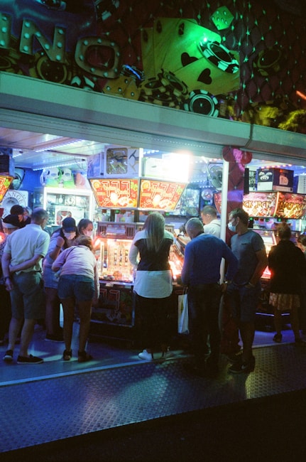 A group of people standing in front of an illuminated amusement arcade, engaging with various gaming machines. The environment is bustling with activity, and colorful lights create a vibrant atmosphere. The ceiling is decorated with large, playful images of dice and other game-related graphics, enhancing the lively mood.