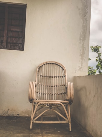 A bamboo chair is positioned against a plain white wall with a small window to the left. The setting appears to be an outdoor terrace or balcony area with a rough, textured wall and a glimpse of greenery on the right side. The scene has a rustic and simple aesthetic.
