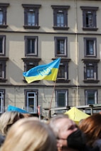 A blue and yellow flag with a trident emblem is prominently displayed on a pole in front of a multi-story building with several windows. The building has a beige facade with gray window frames. Below the flag is a group of people, some of whom are slightly blurred in the foreground.