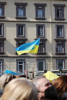 A blue and yellow flag with a trident emblem is prominently displayed on a pole in front of a multi-story building with several windows. The building has a beige facade with gray window frames. Below the flag is a group of people, some of whom are slightly blurred in the foreground.