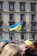 A blue and yellow flag with a trident emblem is prominently displayed on a pole in front of a multi-story building with several windows. The building has a beige facade with gray window frames. Below the flag is a group of people, some of whom are slightly blurred in the foreground.