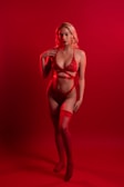 A woman stands confidently against a vibrant red backdrop, wearing red lingerie. The lighting emphasizes the striking color, casting an intense and dramatic tone to the composition. Her pose is poised and self-assured, with one hand resting on her hip and the other touching her shoulder.