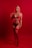 A woman stands confidently against a vibrant red backdrop, wearing red lingerie. The lighting emphasizes the striking color, casting an intense and dramatic tone to the composition. Her pose is poised and self-assured, with one hand resting on her hip and the other touching her shoulder.