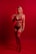 A woman stands confidently against a vibrant red backdrop, wearing red lingerie. The lighting emphasizes the striking color, casting an intense and dramatic tone to the composition. Her pose is poised and self-assured, with one hand resting on her hip and the other touching her shoulder.