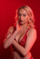 A person with blonde hair poses against a vibrant red background. They are wearing red lingerie and have a calm, confident expression. The lighting emphasizes the warm tones, creating a dramatic effect.