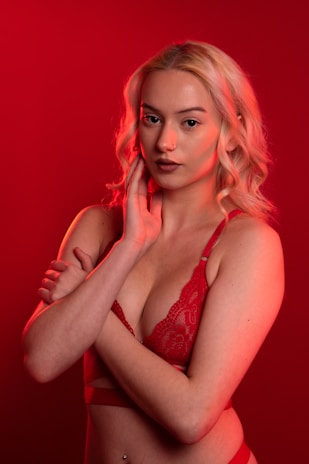 A person with blonde hair poses against a vibrant red background. They are wearing red lingerie and have a calm, confident expression. The lighting emphasizes the warm tones, creating a dramatic effect.