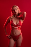 A person is standing against a red background, wearing red lingerie adorned with lace and ribbon details. They have blonde hair and are striking a confident pose with one hand on their hip and the other resting behind their neck.