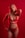 A person is standing against a red background, wearing red lingerie adorned with lace and ribbon details. They have blonde hair and are striking a confident pose with one hand on their hip and the other resting behind their neck.
