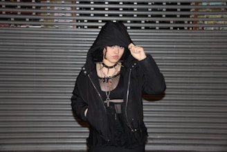 A person wearing a black hooded jacket and a mesh top poses in front of a metal shutter. Their expression is serious, with multiple necklaces and piercings visible, adding to a gothic or alternative style.