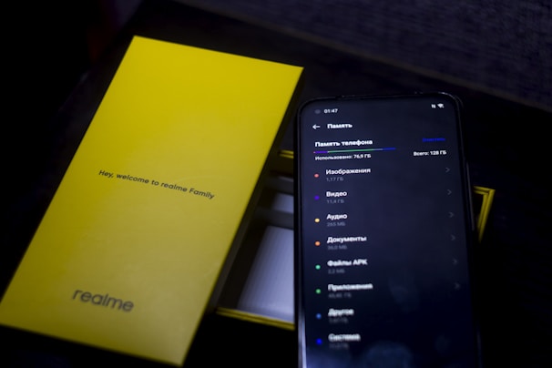 A smartphone displays storage usage information on its screen, showing categories like images, video, and audio files. Next to it, a bright yellow product box with 'Hey, welcome to realme Family' and 'realme' branding stands out.