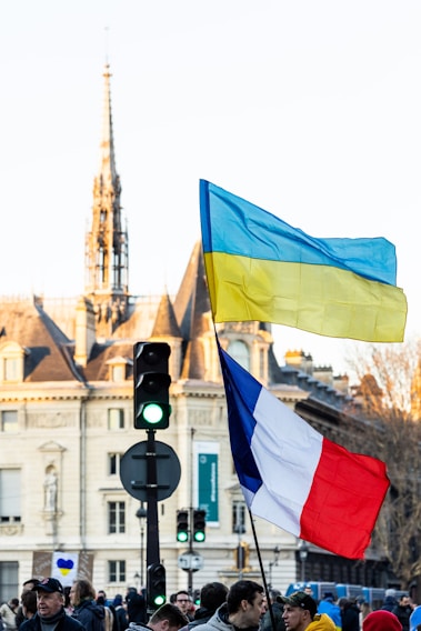 A crowd of people is gathered in a city street, holding two large flags, one Ukrainian and one French. In the background, a historical building with ornate architecture is visible. The scene also includes a traffic light showing green.