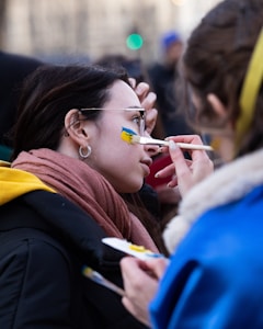 A person is having a flag painted on their cheek by another individual using a paintbrush and palette. The scene likely takes place outdoors with other blurred figures in the background. The subject is wearing a scarf and jacket.