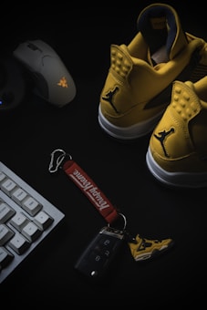 A pair of yellow sneakers with a distinctive logo on a dark background, accompanied by a gaming mouse on the left side and a desktop keyboard partially visible at the bottom. A set of car keys with a keychain featuring miniature sneaker design and branding are placed in the center.