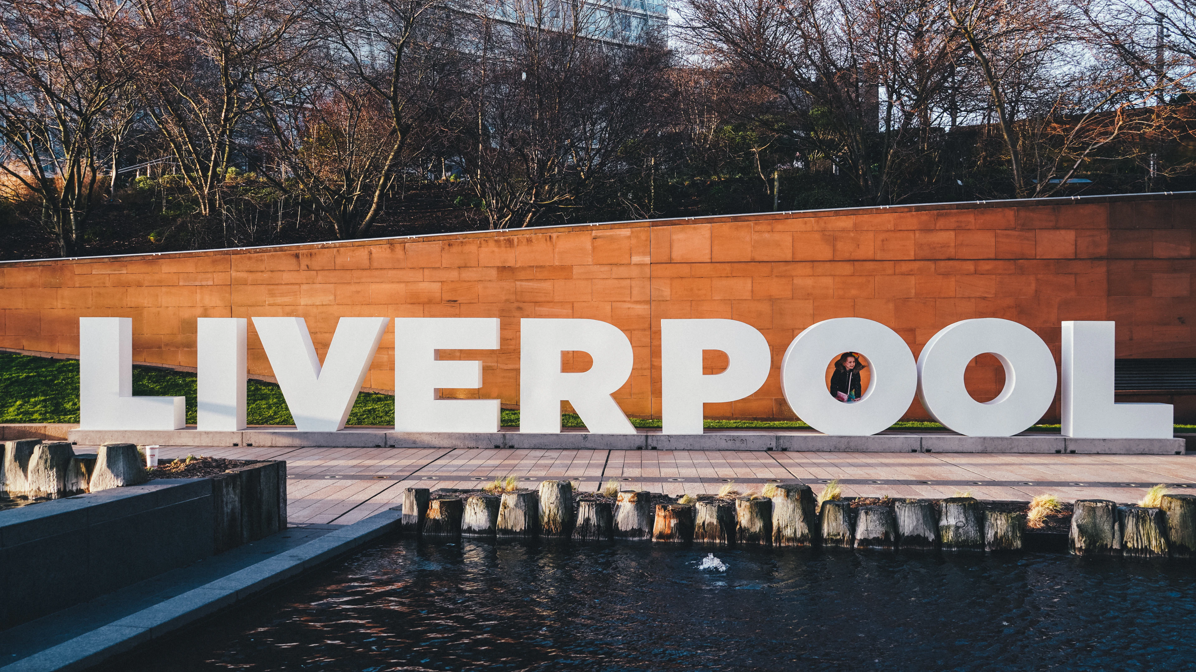 The large 'LIVERPOOL' letter sculpture at the waterfront, lit warmly at dusk with a figure silhouetted between the letters.