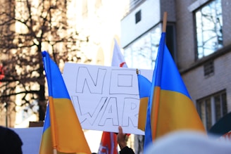 A group of people are gathered in a protest holding Ukrainian flags and a sign that says 'NO WAR'. The setting appears to be an urban environment with buildings and trees in the background.