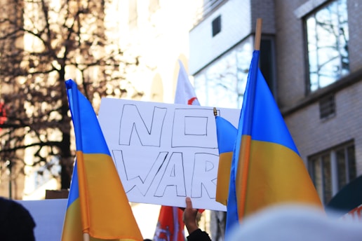 A group of people are gathered in a protest holding Ukrainian flags and a sign that says 'NO WAR'. The setting appears to be an urban environment with buildings and trees in the background.