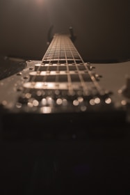 A close-up perspective of an electric guitar, focusing on the strings and fretboard as they lead up towards the headstock. The lighting creates a dramatic effect, with highlights on the strings and a soft, blurred background.
