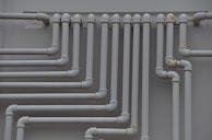 A series of parallel white pipes with multiple bends and joints is arranged on a light gray wall. The pipes display slight rust and wear in certain areas, indicating exposure to elements over time.
