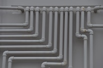 A series of parallel white pipes with multiple bends and joints is arranged on a light gray wall. The pipes display slight rust and wear in certain areas, indicating exposure to elements over time.