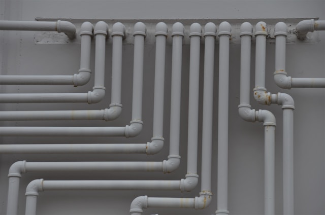 A series of parallel white pipes with multiple bends and joints is arranged on a light gray wall. The pipes display slight rust and wear in certain areas, indicating exposure to elements over time.