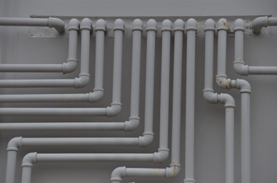 A series of parallel white pipes with multiple bends and joints is arranged on a light gray wall. The pipes display slight rust and wear in certain areas, indicating exposure to elements over time.