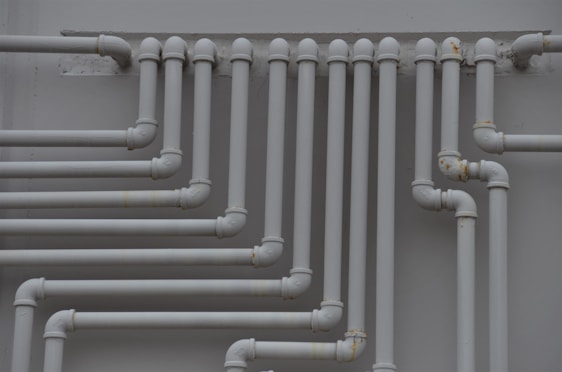 A series of parallel white pipes with multiple bends and joints is arranged on a light gray wall. The pipes display slight rust and wear in certain areas, indicating exposure to elements over time.
