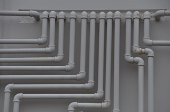 A series of parallel white pipes with multiple bends and joints is arranged on a light gray wall. The pipes display slight rust and wear in certain areas, indicating exposure to elements over time.