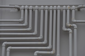 A series of parallel white pipes with multiple bends and joints is arranged on a light gray wall. The pipes display slight rust and wear in certain areas, indicating exposure to elements over time.