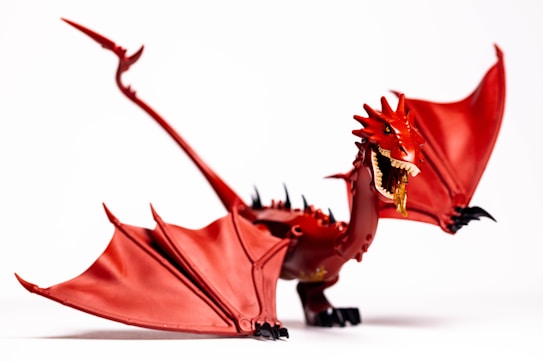 A detailed model of a red dragon with large wings, sharp claws, and an open mouth displaying its teeth and what appears to be flames emerging. The dragon is posed dynamically, highlighting its spiked back and long tail, set against a plain white background.