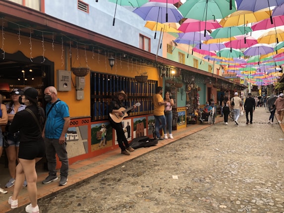 A vibrant street with colorful umbrellas hanging overhead. People are walking and some are standing by a building with bright yellow and blue storefronts. A musician plays guitar, adding to the lively atmosphere. Various shops showcase artistic decorations and goods.