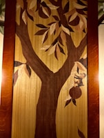 A wooden wall art piece depicts a large tree with branches and leaves. A small monkey is seen climbing the tree using one of its branches, with a piece of fruit hanging nearby. The art is framed by a wooden border.