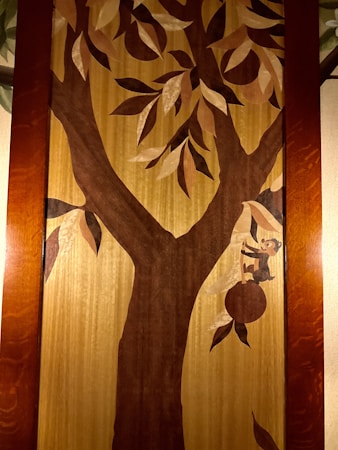 A wooden wall art piece depicts a large tree with branches and leaves. A small monkey is seen climbing the tree using one of its branches, with a piece of fruit hanging nearby. The art is framed by a wooden border.