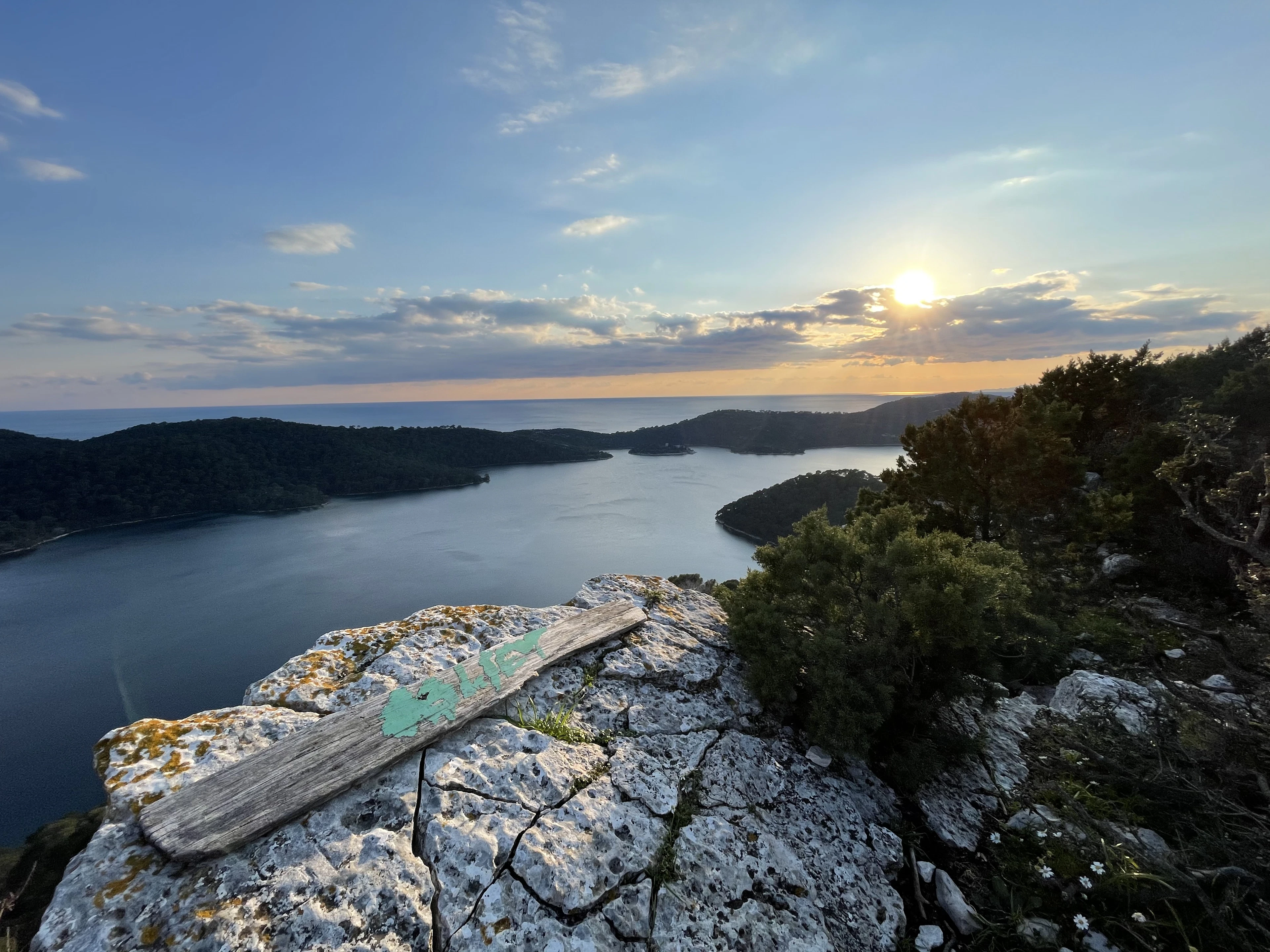 Sunset over the salt lakes of Mljet National Park seen from the ridge above, a weathered wooden trail marker lying on the white limestone in the foreground and wooded headlands folding into the water.