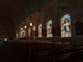 A dimly lit church interior featuring rows of wooden pews and several beautiful stained glass windows on one side. The stained glass depicts various religious scenes, casting colorful light patterns into the room. Hanging light fixtures illuminate the area softly, contributing to the serene ambiance. At the far end, an altar can be seen.