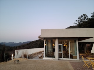 A modern, square-shaped house with large windows is set against a backdrop of rolling hills and trees. The outdoor area features a small patio with a couple of chairs and tables. Soft lighting from within the house creates a warm atmosphere, and the sky is clear during what appears to be dawn or dusk.