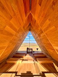 A wooden A-frame interior structure with a large triangular window at the far end. The window offers a view of rolling hills and a blue sky. Inside the structure, there are tiered wooden benches leading up to the window. Natural light fills the space, highlighting the warm tones of the wood.