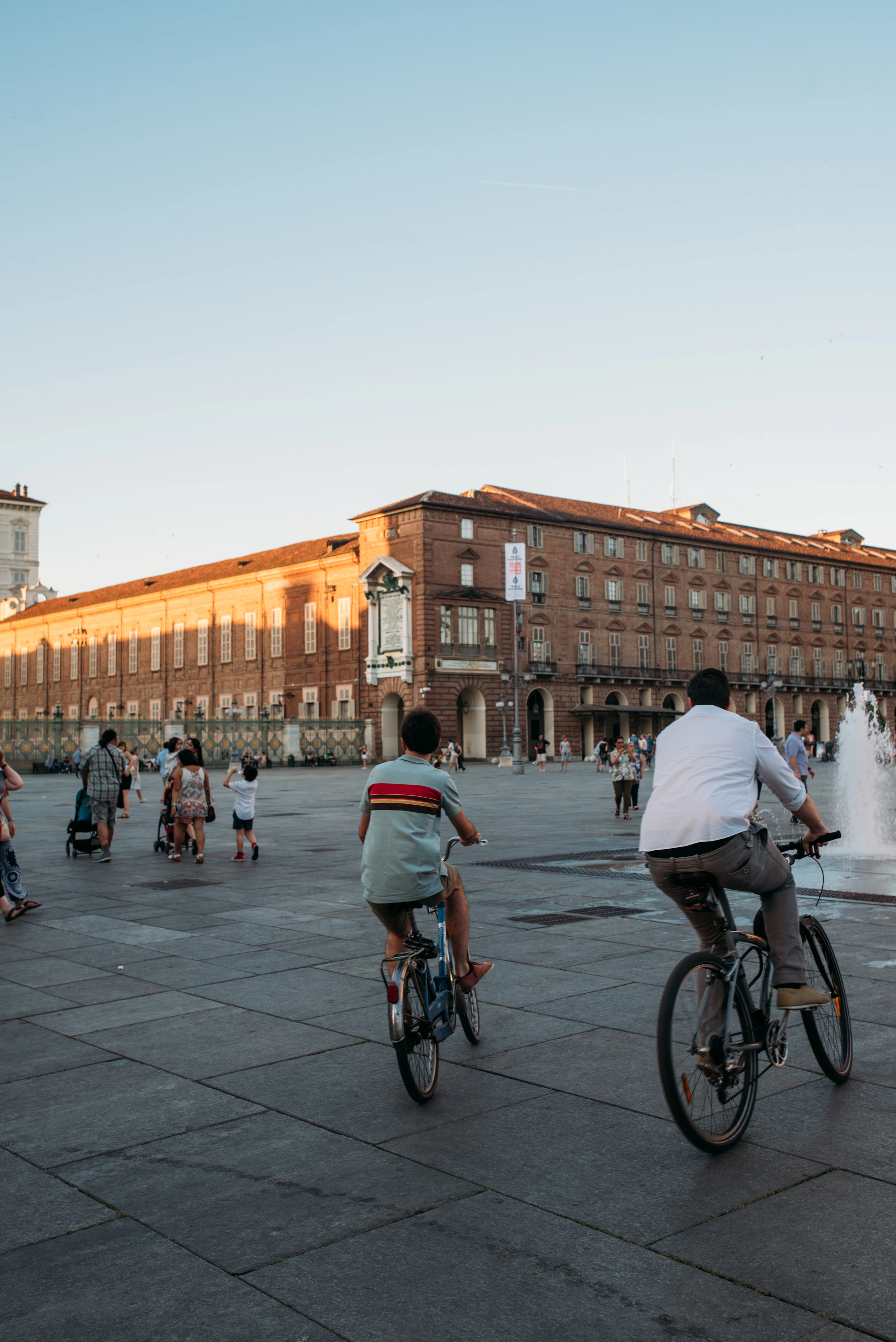 Cyclists ride across a grand piazza past a fountain and arcaded palazzo at golden hour.