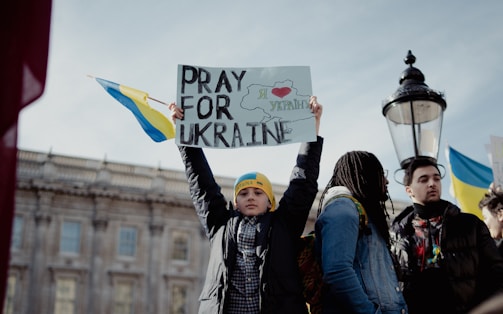 A group of people gathered with a young person holding a sign that reads 'Pray for Ukraine'. The sign includes the outline of Ukraine and a heart. Some individuals are dressed warmly, and Ukrainian flags are also present. The setting appears to be outdoors, near a building with classical architecture.