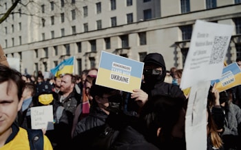 A crowd of protesters holds various signs and flags, including a prominent one reading 'Hongkongers Stand with Ukraine.' The group is gathered in front of a large building, possibly governmental, under a clear sky. Many in the crowd are wearing masks and holding up flags that feature blue and yellow colors, signifying support for Ukraine.