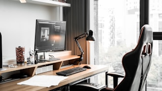 A modern workspace featuring a computer monitor displaying architectural software, a sleek black desk lamp, a gaming-style office chair, and various desk items including a keyboard, mouse, drink container, and paperwork. The desk is positioned by a large window with a view of a cityscape.
