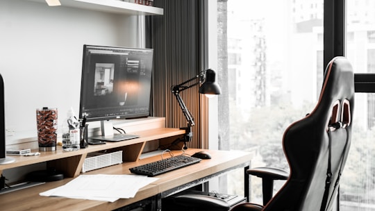 A modern workspace featuring a computer monitor displaying architectural software, a sleek black desk lamp, a gaming-style office chair, and various desk items including a keyboard, mouse, drink container, and paperwork. The desk is positioned by a large window with a view of a cityscape.