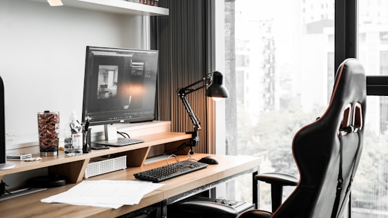 A modern workspace featuring a computer monitor displaying architectural software, a sleek black desk lamp, a gaming-style office chair, and various desk items including a keyboard, mouse, drink container, and paperwork. The desk is positioned by a large window with a view of a cityscape.