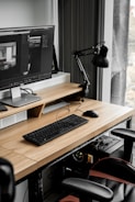 A modern workspace with a large computer monitor displaying 3D modeling software, set on a wooden desk. A keyboard and mouse are placed neatly in front of the monitor. The scene includes a black adjustable desk lamp on the right and a visible computer tower underneath the desk. The workspace is by a window with sheer curtains, allowing natural light to enter.