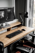 A modern workspace with a large computer monitor displaying 3D modeling software, set on a wooden desk. A keyboard and mouse are placed neatly in front of the monitor. The scene includes a black adjustable desk lamp on the right and a visible computer tower underneath the desk. The workspace is by a window with sheer curtains, allowing natural light to enter.