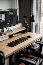 A modern workspace with a large computer monitor displaying 3D modeling software, set on a wooden desk. A keyboard and mouse are placed neatly in front of the monitor. The scene includes a black adjustable desk lamp on the right and a visible computer tower underneath the desk. The workspace is by a window with sheer curtains, allowing natural light to enter.