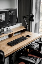 A modern workspace with a large computer monitor displaying 3D modeling software, set on a wooden desk. A keyboard and mouse are placed neatly in front of the monitor. The scene includes a black adjustable desk lamp on the right and a visible computer tower underneath the desk. The workspace is by a window with sheer curtains, allowing natural light to enter.