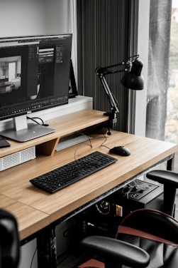 A modern workspace with a large computer monitor displaying 3D modeling software, set on a wooden desk. A keyboard and mouse are placed neatly in front of the monitor. The scene includes a black adjustable desk lamp on the right and a visible computer tower underneath the desk. The workspace is by a window with sheer curtains, allowing natural light to enter.