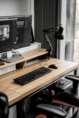 A modern workspace with a large computer monitor displaying 3D modeling software, set on a wooden desk. A keyboard and mouse are placed neatly in front of the monitor. The scene includes a black adjustable desk lamp on the right and a visible computer tower underneath the desk. The workspace is by a window with sheer curtains, allowing natural light to enter.