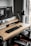 A modern workspace with a large computer monitor displaying 3D modeling software, set on a wooden desk. A keyboard and mouse are placed neatly in front of the monitor. The scene includes a black adjustable desk lamp on the right and a visible computer tower underneath the desk. The workspace is by a window with sheer curtains, allowing natural light to enter.